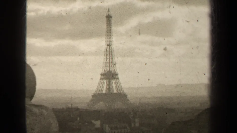 Vintage 8mm Paris frame showing the Eiffel Tower below an overcast sky, with faint city rooftops and film wear visible.