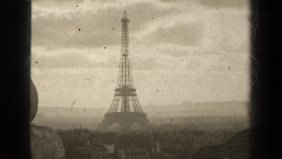 1947 8mm Paris view of the Eiffel Tower through a window, with soft sepia tone, cloudy sky, and faint rooftops below.