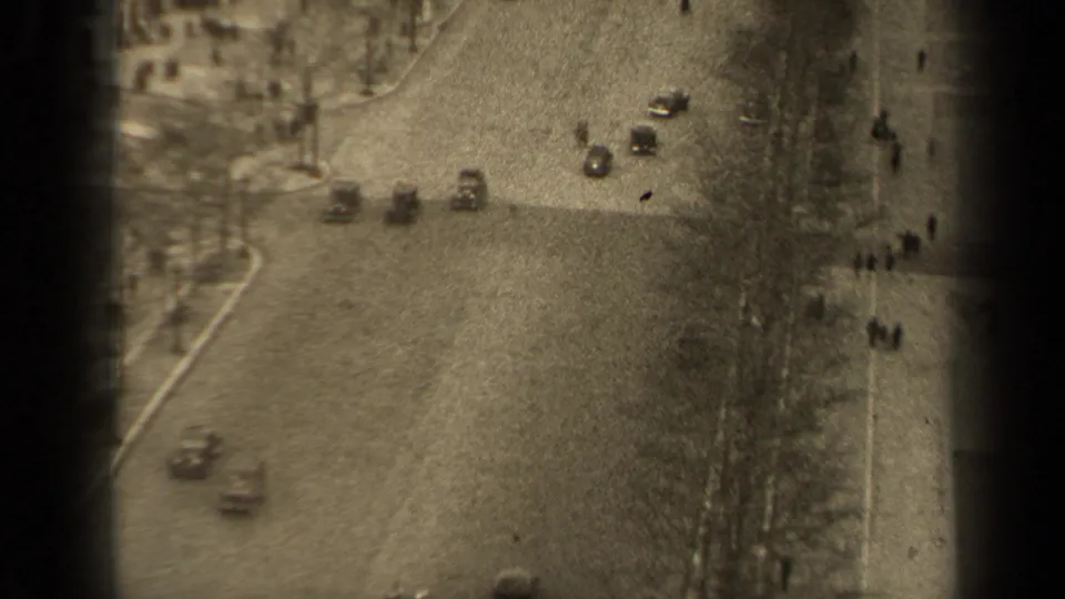 High-angle Paris street frame showing several classic cars, pedestrians, roadside trees, and heavy 8mm grain.