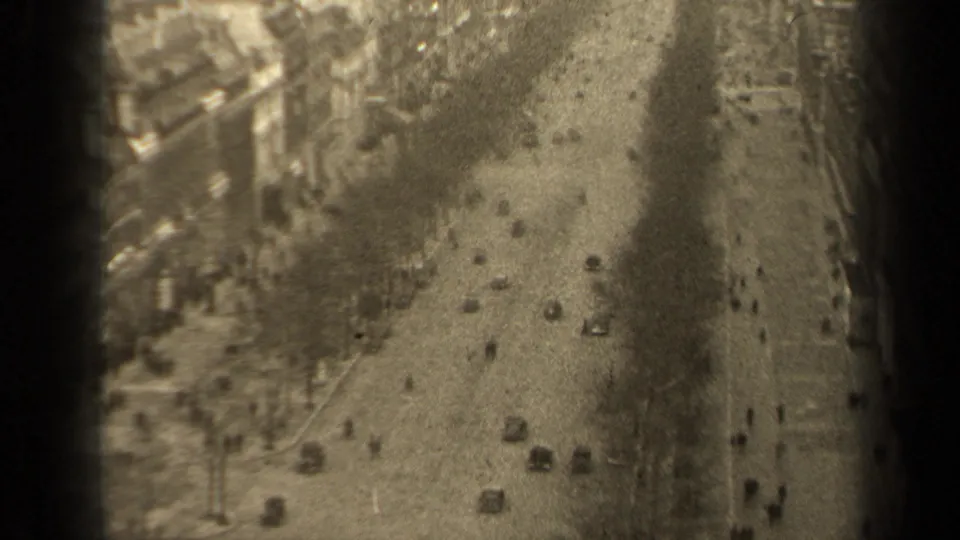 1947 Paris boulevard seen from above, with small cars, pedestrians, bare trees, and building fronts in soft black-and-white 8mm texture.