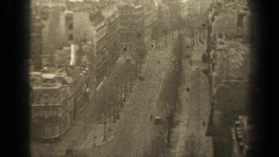 High-angle 1947 Paris boulevard view with multi-story buildings, rows of trees, pedestrians and a few cars moving through the street.