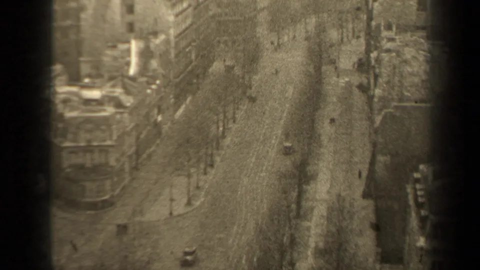 Grainy 8mm frame of a 1947 Paris boulevard seen from above, with vintage cars, pedestrians, sidewalks, trees and dense urban buildings.