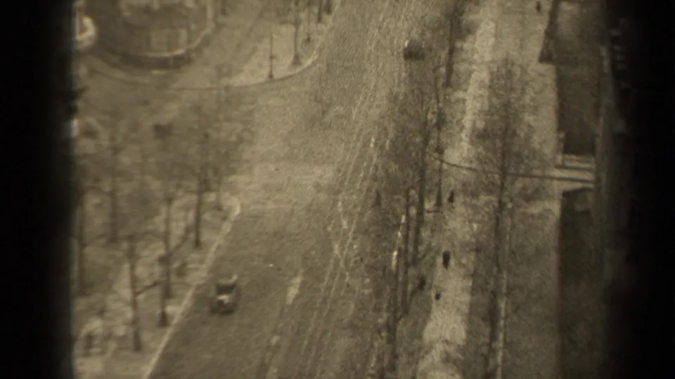 1947 Paris home-movie frame looking down on a sparse boulevard with bare trees, sidewalks, distant pedestrians, and one small car.