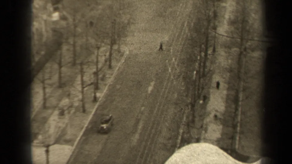 Grainy 8mm frame of a Paris boulevard from above, showing a small car, scattered pedestrians, bare trees, and a bright lower window ledge.