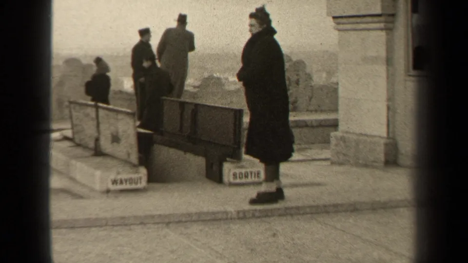 Late-1940s Paris 8mm frame with a woman watching near a bilingual exit as other visitors move through.