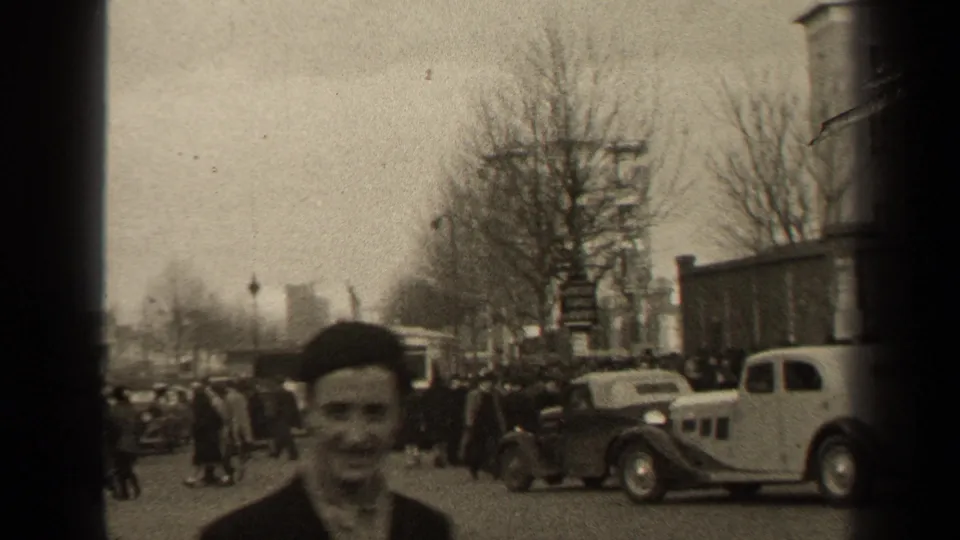 A smiling man in a dark cap appears close to the camera on a 1947 Paris street, with cars and pedestrians behind him.