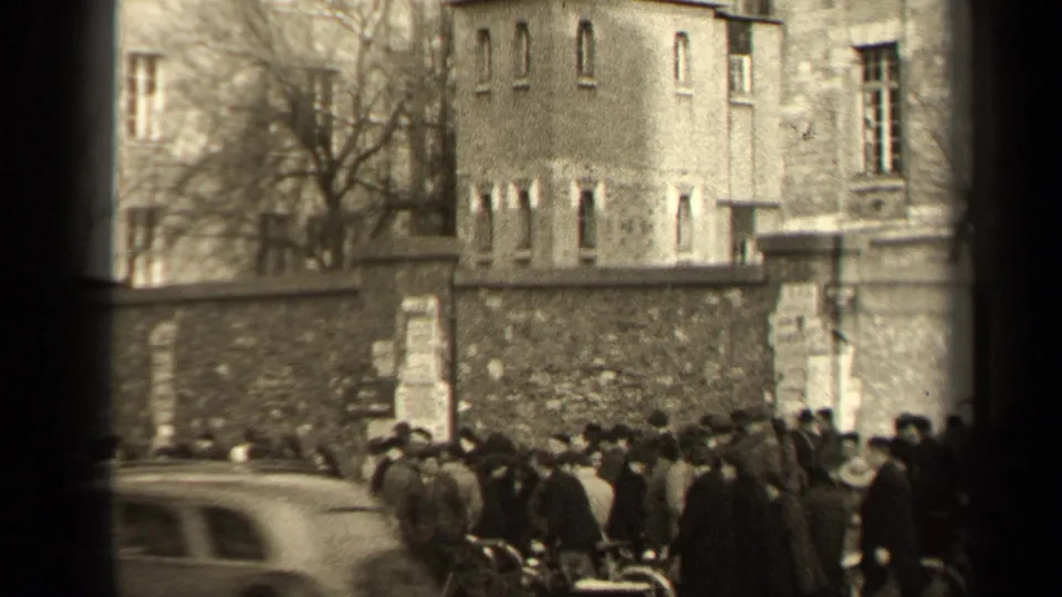 1947 Paris street crowd with bicycles by a stone wall and a blurred car crossing the lower frame.