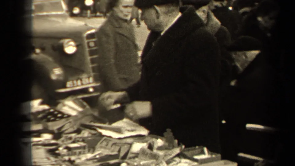 1947 Paris sidewalk close view of pedestrians gathered around a vendor table with papers and small goods, with vintage cars nearby.