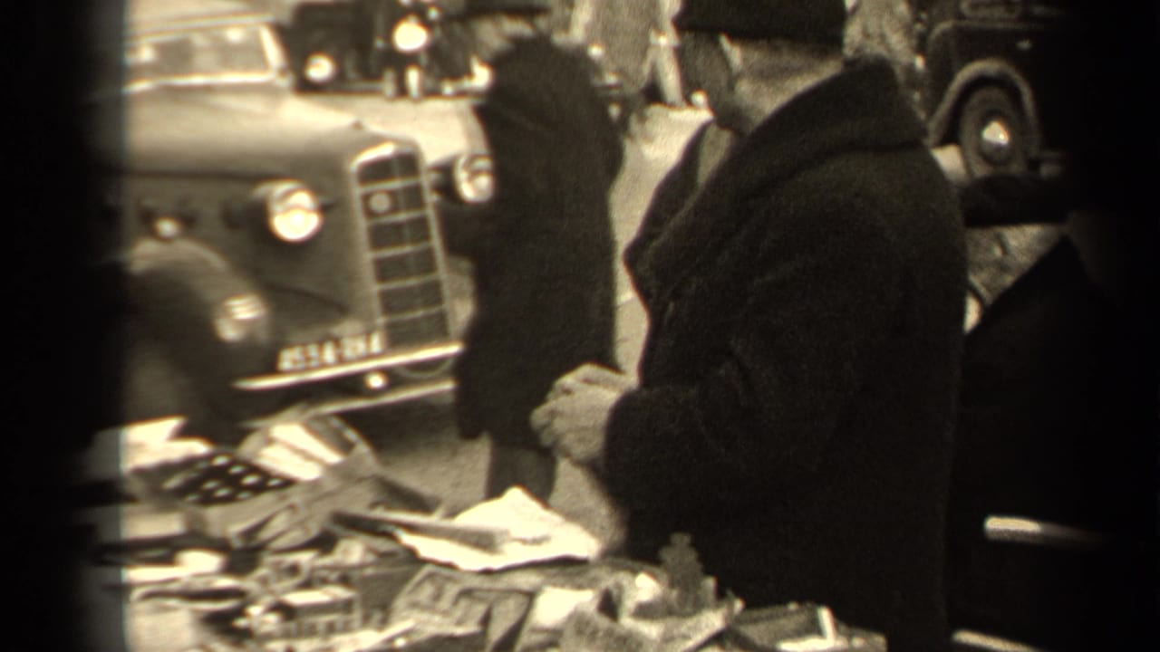 Man in a dark coat browsing a street market table with mirrors and papers beside parked cars.