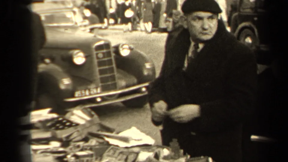 In this 1947 Paris market frame, a man in a dark cap stands beside a table of secondhand objects with a period car in the background.