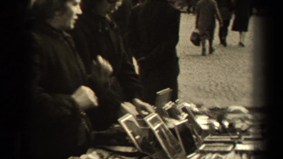 Women in period coats browse a Paris outdoor market table with small framed items and trinkets visible in the foreground.