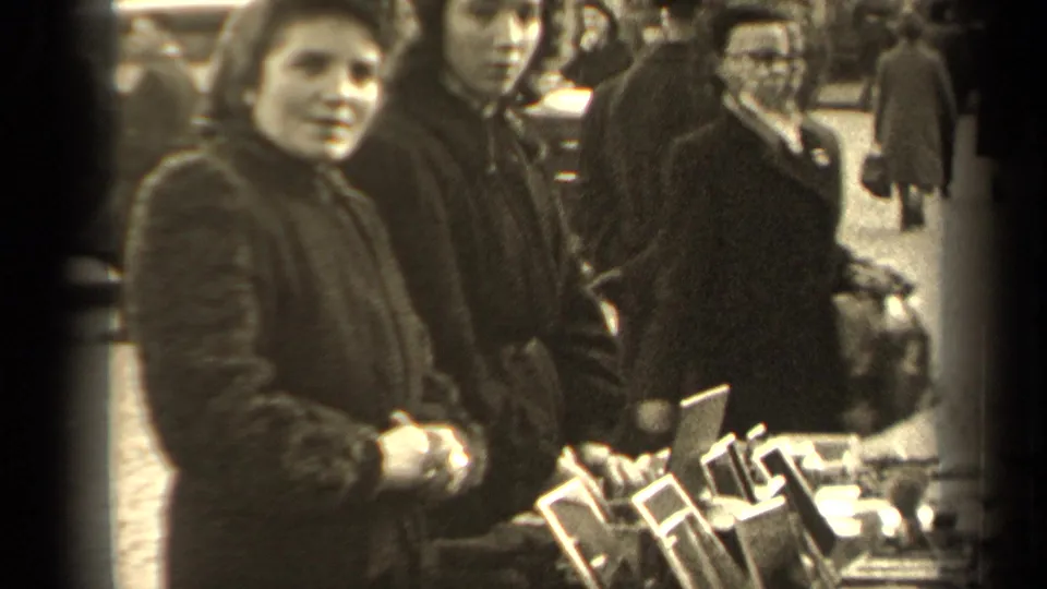 A woman at a 1947 Paris outdoor market looks toward the camera near a tabletop display while other people stand behind her.