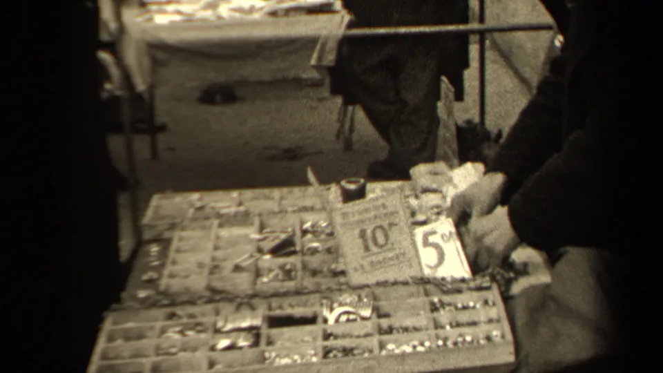 Close market-stall view in Paris with hands browsing a small-goods tray and handwritten price cards.
