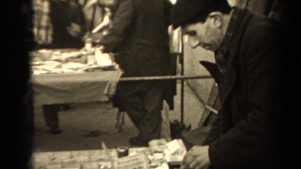 A man in a cap leans toward a Paris market stall as hands handle small wares near price cards.