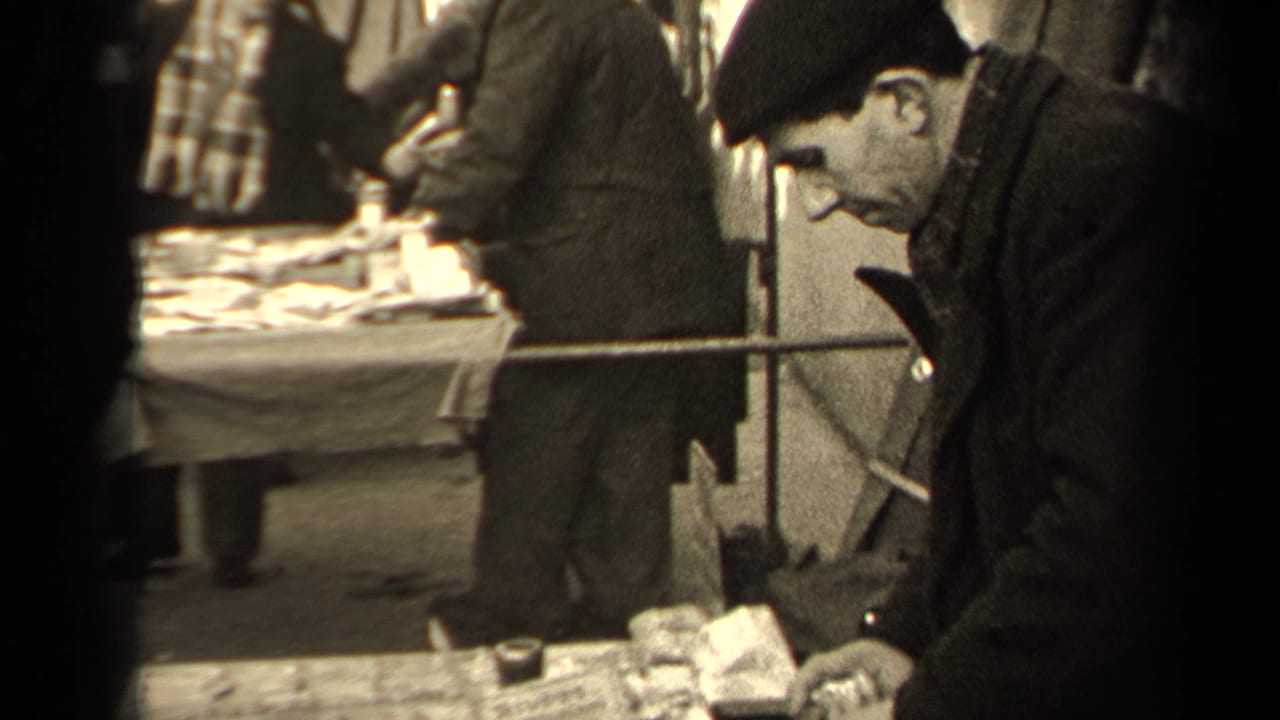 Man in a dark coat and cap leans over a market table as pedestrians pass behind him.