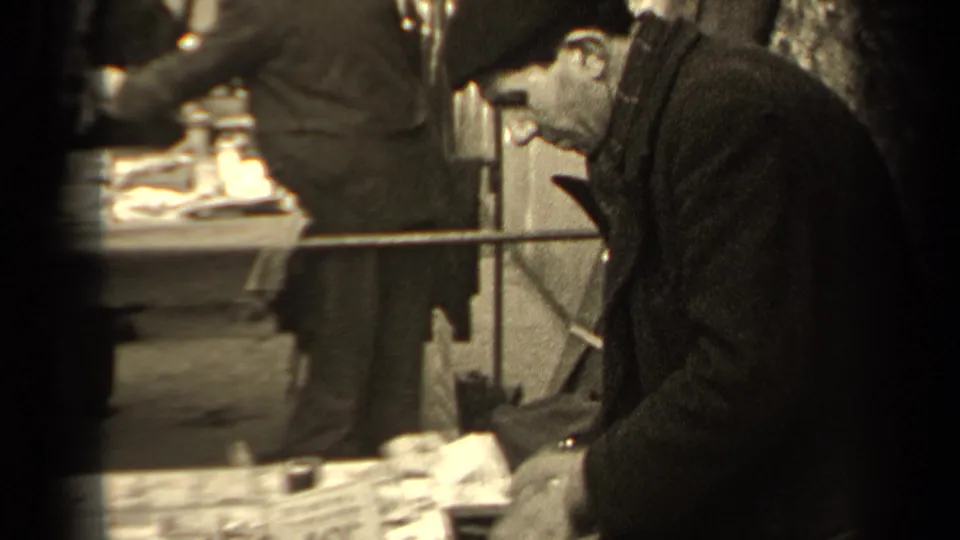 A 1947 Paris street-market view shows a man in a cap beside a table of goods, with pedestrians and stall materials behind him.