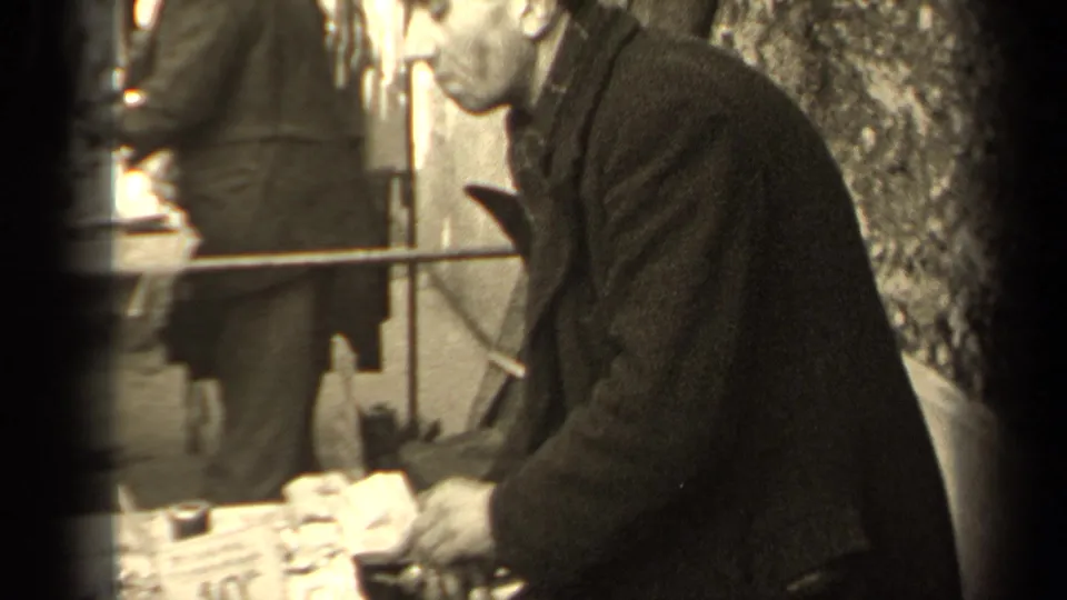 The home-movie camera passes close to a man in a dark coat at a Paris market table, with street figures and a tree trunk nearby.