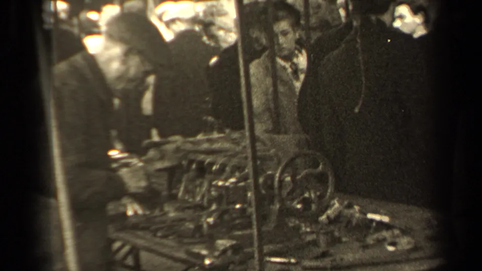 Paris market shoppers stand around a table of tools and metal mechanical parts, with a woman in plaid visible behind the display.