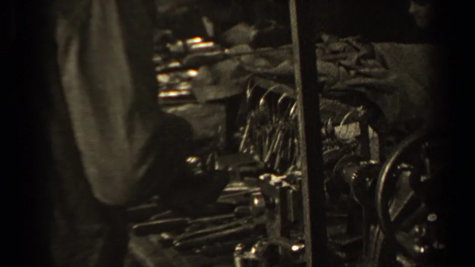 1947 Paris home-movie frame partly obscured by a standing figure, with reflective metal pieces and a workbench still visible.