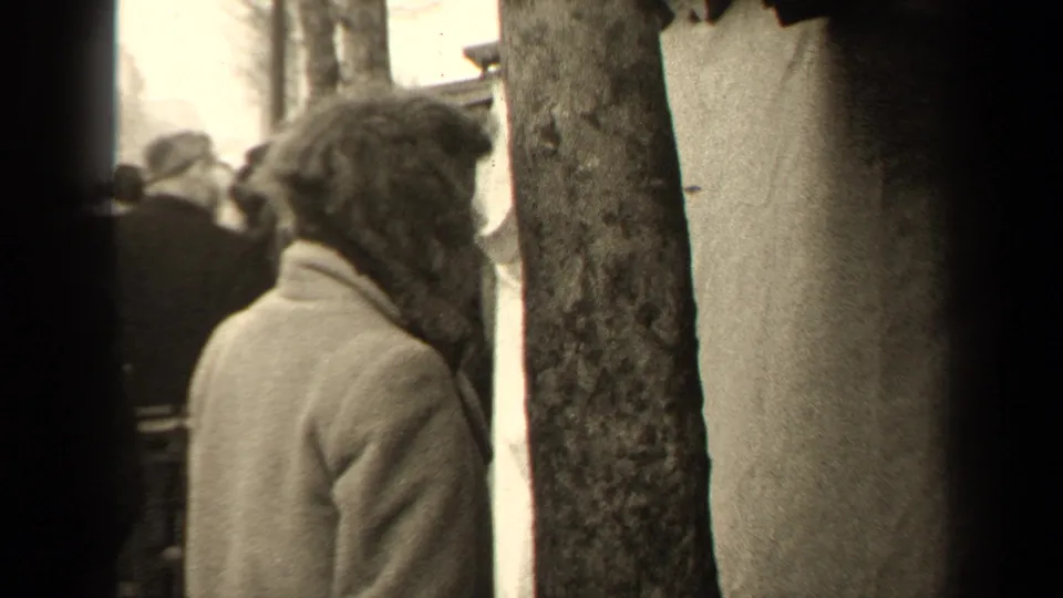 Paris 1947 8mm frame showing a person in a heavy coat near a tree trunk, with several people gathered in the background.