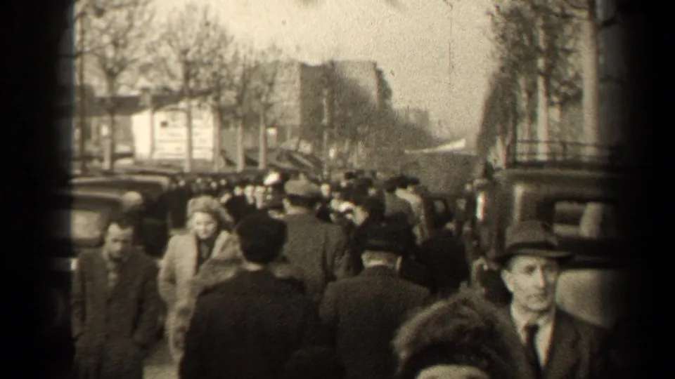 A busy Paris street in 1947 appears crowded with pedestrians in coats and hats, with older cars and bare trees behind them.