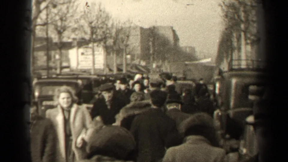 In the 1947 Paris street scene, a woman in a light coat walks toward the camera as the sidewalk remains crowded behind her.