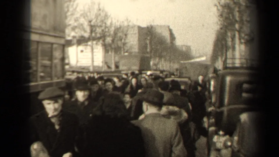 Crowded 1947 Paris sidewalk with several faces near camera, winter clothing, vintage cars, and bare boulevard trees in view.