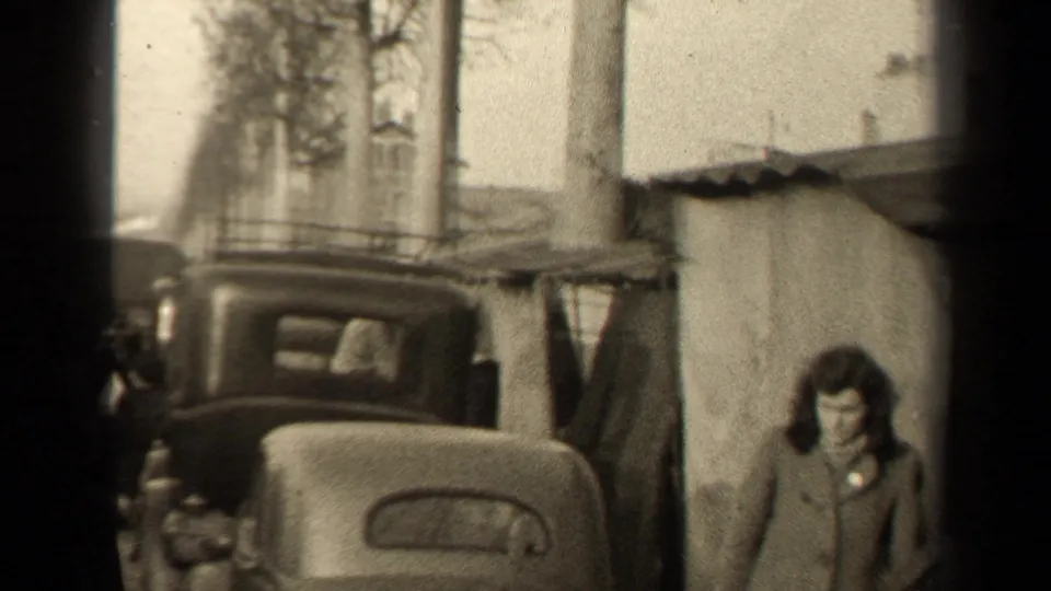 1947 Paris street frame showing an older car, bare trees, a tarp-covered shelter, and a woman in a heavy coat.