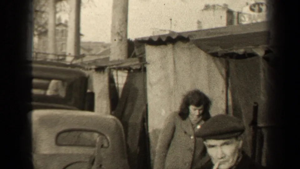 1947 Paris sidewalk scene with a woman by a tarp-covered structure, an older car, and a man in a flat cap crossing the foreground.