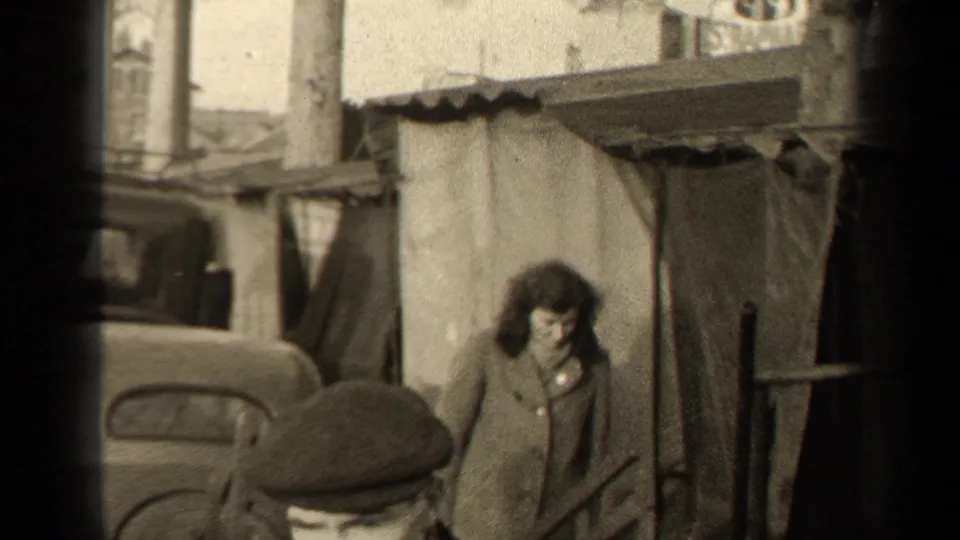 1947 Paris street view with a man in a flat cap in the foreground, a woman in a coat, older cars, and a rough covered shelter.