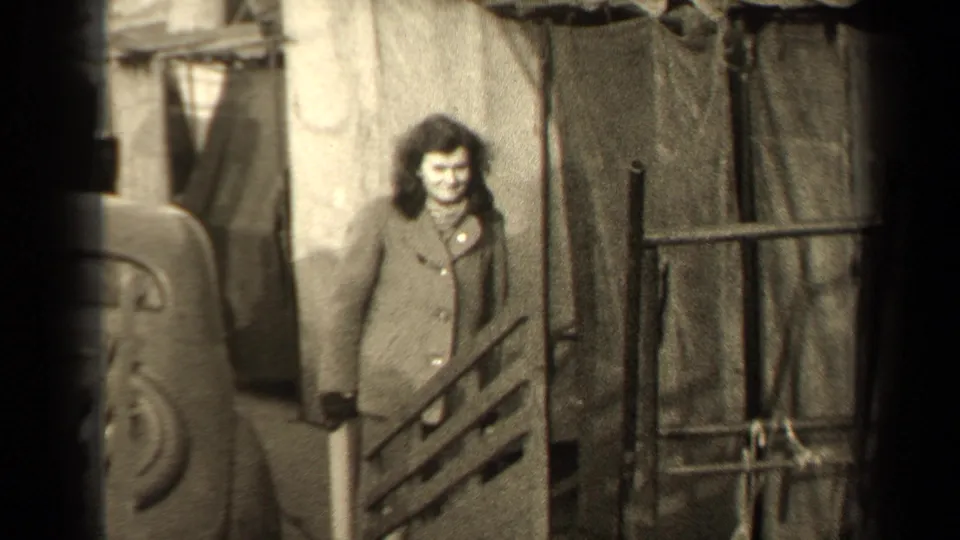 1947 Paris home-movie view of a woman in a buttoned coat near railings, a car, and canvas-covered street stalls or shelters.