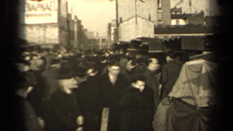 1947 Paris crowd scene with pedestrians in coats and hats passing close to covered sidewalk stalls on a busy urban street.
