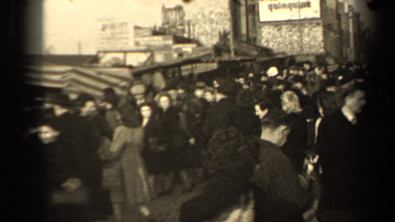 People in coats and hats move through a crowded street beside awnings and shopfronts.