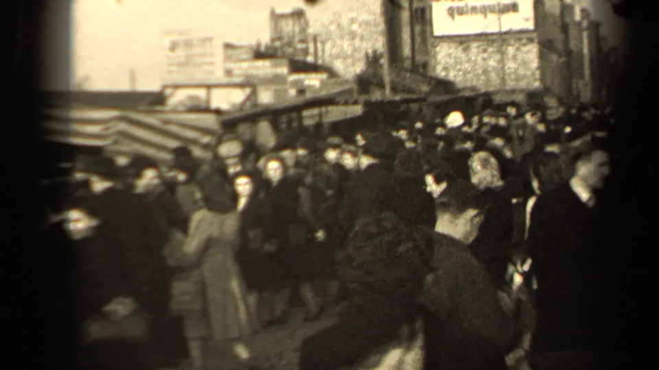 Mid-century Paris pedestrians pass through a tightly packed street scene with awnings, coats, hats, and partially readable signage.