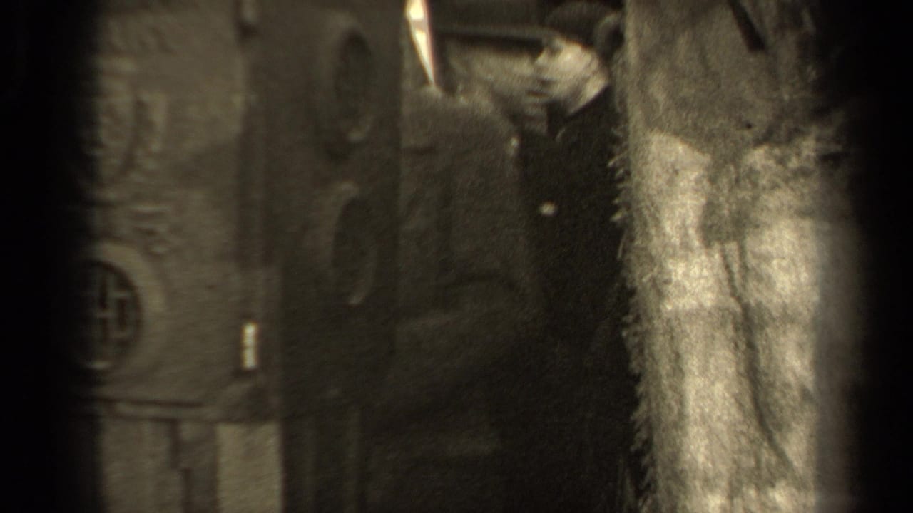 People in heavy coats stand near a wall while a light-colored garment hangs in the foreground.