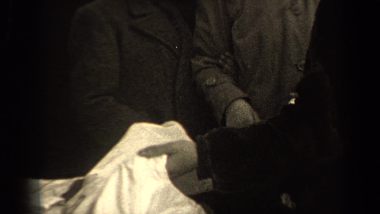 In 1947 Paris home-movie footage, adults in dark coats handle a light-colored shirt under low indoor light.