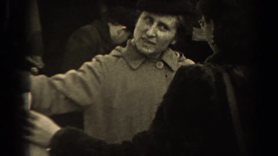 In the Paris market footage, a woman in a buttoned coat appears to speak or gesture while a garment is held in front of her.