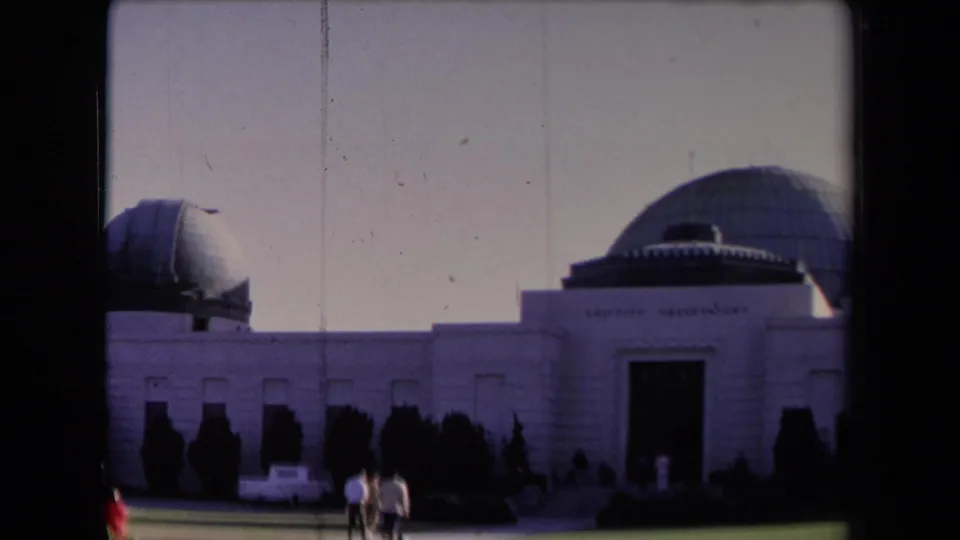 1967 Los Angeles home-movie frame showing visitors approaching the Griffith Observatory facade with domes and a parked white car.