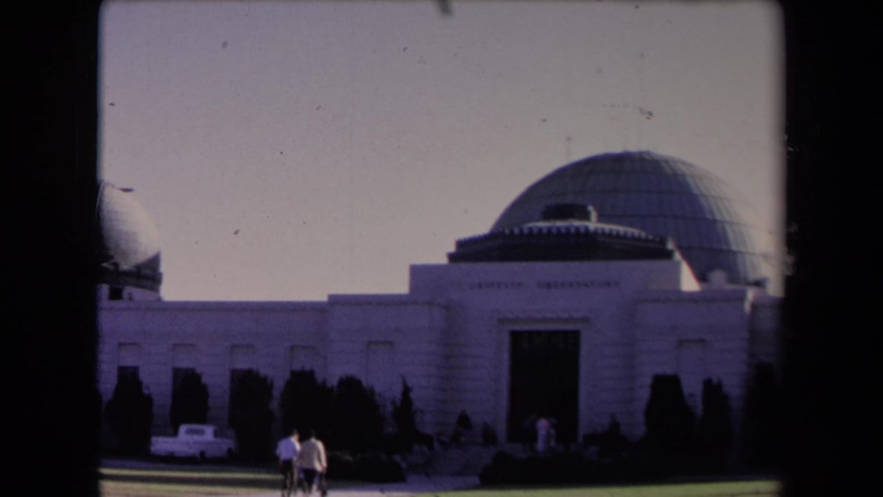 1967 Los Angeles frame of Griffith Observatory with grey domes, dark shrubs, a white car, and visitors moving across the foreground.