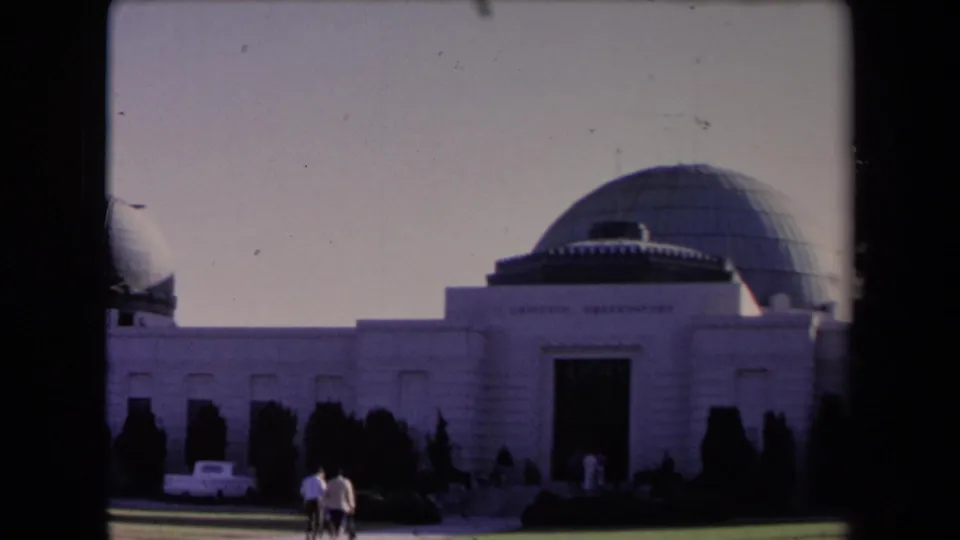 1967 Los Angeles frame of Griffith Observatory with grey domes, dark shrubs, a white car, and visitors moving across the foreground.