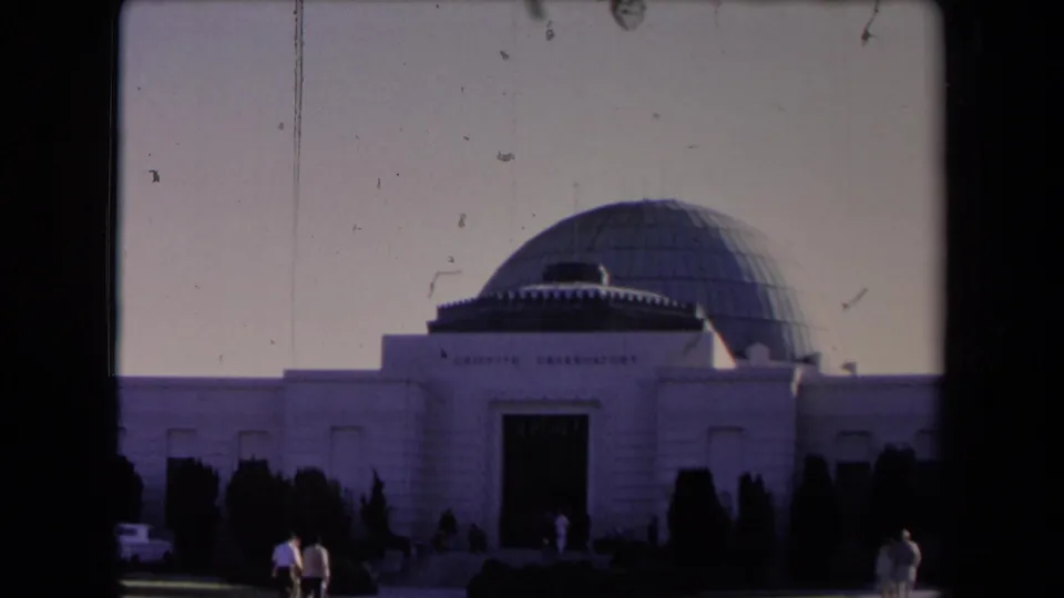 1967 home-movie frame of Griffith Observatory with pedestrians at the lawn edge, dark landscaping, and the grey dome above the white facade.