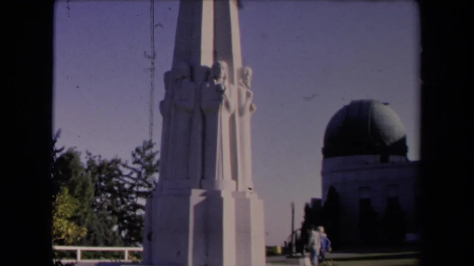 1967 home-movie frame of Griffith Observatory in Los Angeles, with a tall sculpted pillar, dome, trees, railing, and distant visitors.