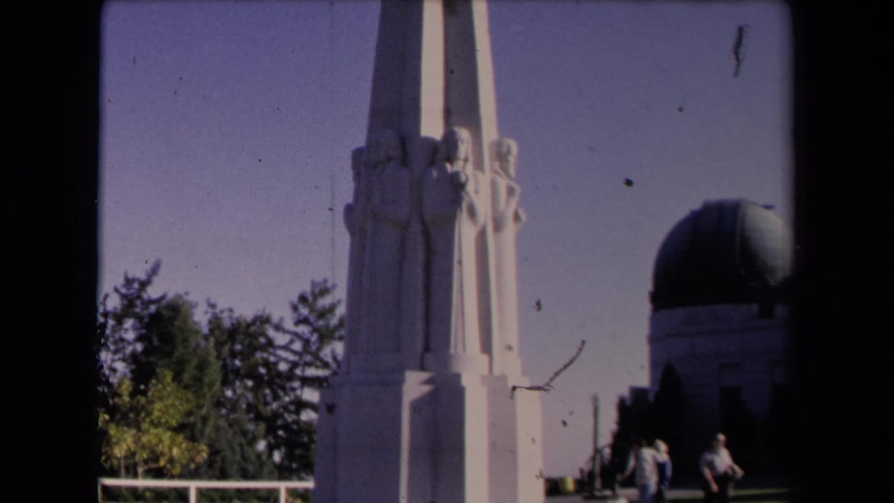 White monument with carved figures in front of the Griffith Observatory dome and visitors.