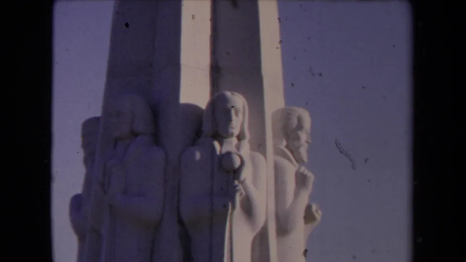 Close 1967 8mm frame of sculpted figures on a tall white pillar at Griffith Observatory in Los Angeles, with the sky behind them.