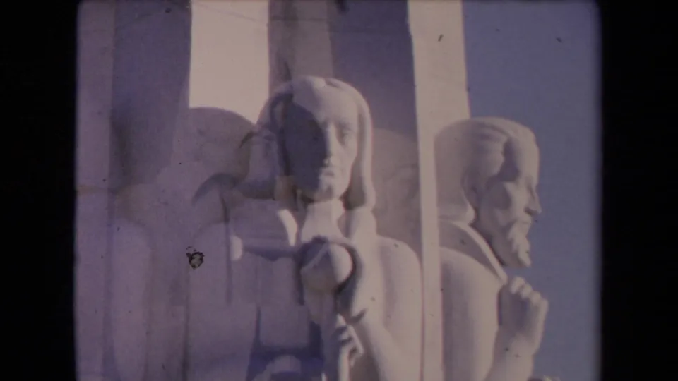 1967 Los Angeles 8mm frame with soft exposure on white stone male figures, showing carved faces, hands, and monument panels.