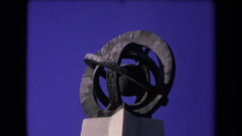 1967 Los Angeles 8mm still showing a dark metal armillary sphere sculpture mounted on a pale pedestal under blue sky.