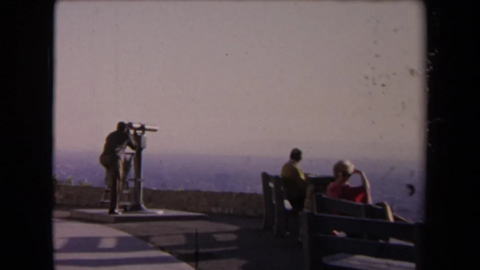 At a Los Angeles vista point in 1967, a person uses a coin-operated telescope while two adults sit on a bench looking toward the hazy cityscape.