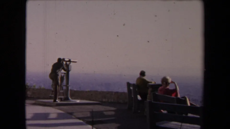 1967 Los Angeles vista point with a coin-operated telescope, seated adults on benches, a low overlook wall, and a hazy city view beyond.