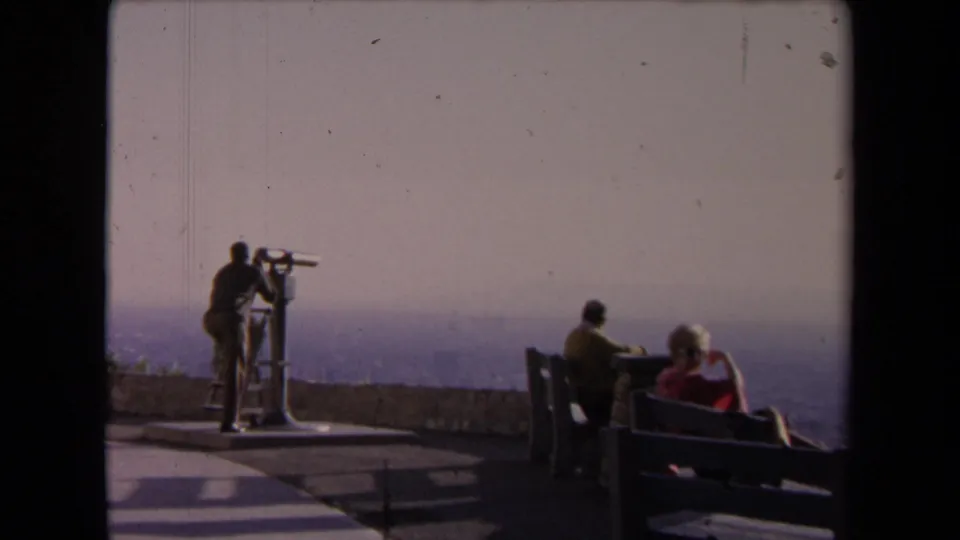 1967 Los Angeles home-movie frame showing visitors on an observatory deck, one using a coin telescope while others sit on benches.