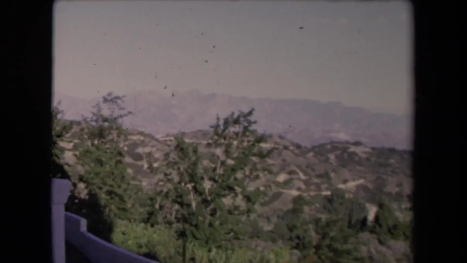 1967 Los Angeles home-movie landscape frame showing scrubby hills, trees, distant mountains, and the edge of a white terrace wall.
